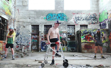 [Image Description: Black and white photo is of Luke, Dan, and Julio, standing apart, in a big abandoned concrete room with graffiti on the walls and huge damaged windows. Luke (white) is standing on the left, he has long flowing blonde hair, arm tattoos, and is wearing a 5 panel hat, a Mars Attacks t-shirt, and black jean shorts on. Dan (white) is in the centre sitting on his silver walker, he has short blonde hair and is wearing a trucker hat, round glasses, is shirtless, and has black shorts and boots on. Julio (white) is on the right, he’s got long dark brown hair, arm tattoos, and is wearing a black 5 panel hat, t-shirt, a sleeveless blue denim jean jacket with patches on, and has black jeans shorts on.]